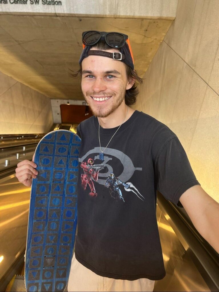 Young man with a beard stands on an escalator, smiling at the camera while holding a blue skateboard and wearing a black graphic t-shirt, cap, and sunglasses on his head.