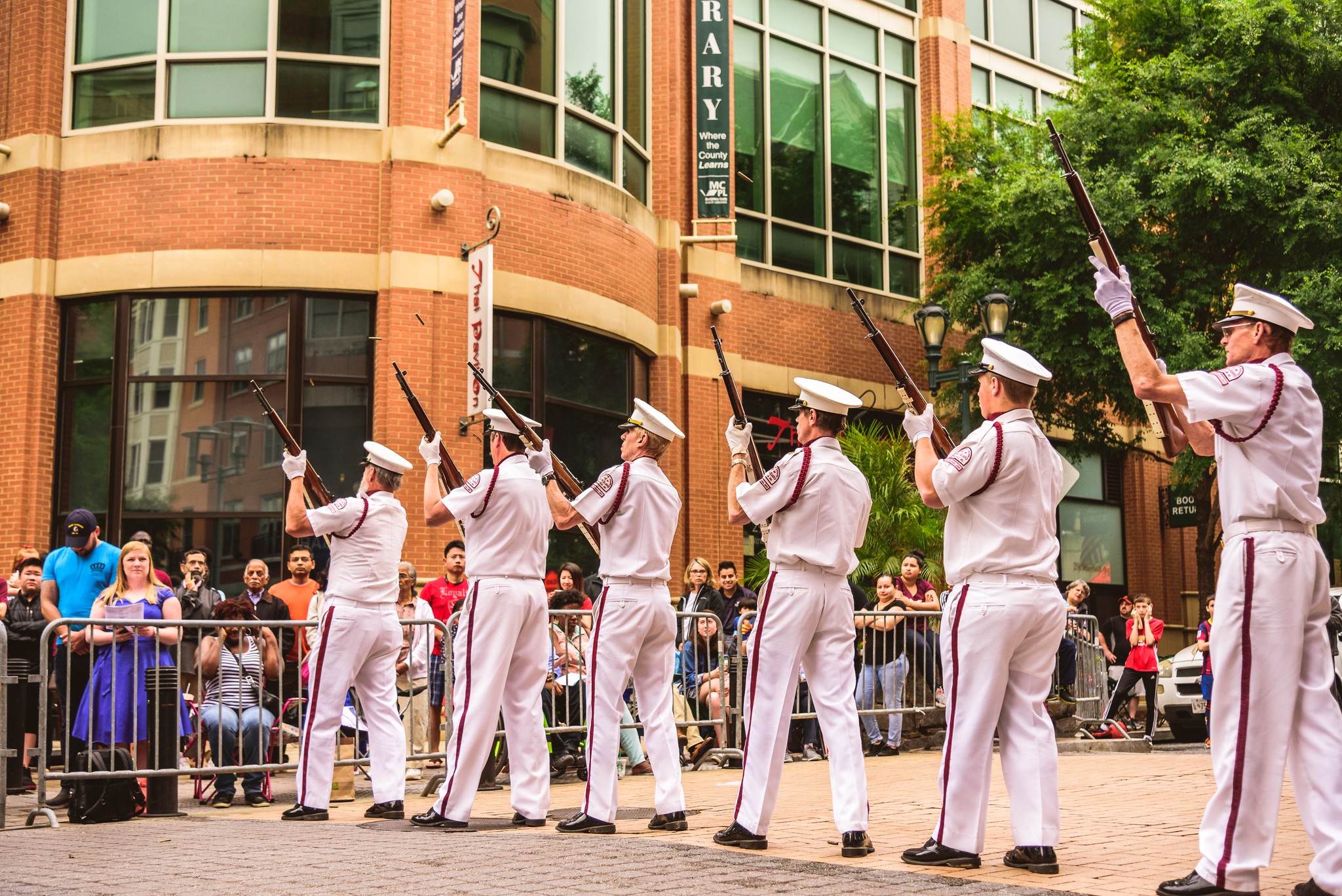Six uniformed military personnel perform a rifle drill in front of a crowd on a city street, with a brick building and trees in the background.