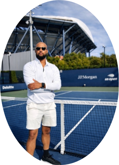 A man wearing a white shirt and shorts stands on a tennis court near the net, with stadium seating and US Open signage in the background.