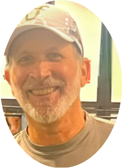 Older man with a gray beard and mustache wearing a light-colored cap and shirt, smiling indoors with blurred background.