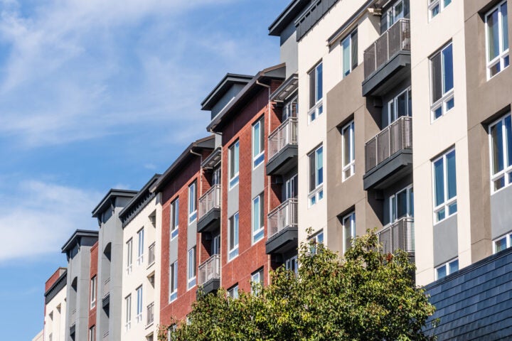 Modern multi-story apartment building with red, gray, and beige facades, featuring balconies and large windows, under a partly cloudy sky with a tree in the foreground.