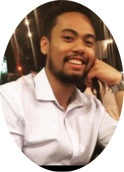 A man with short dark hair and a beard, wearing a white collared shirt, sits at a table and smiles at the camera with his head resting on his hand.