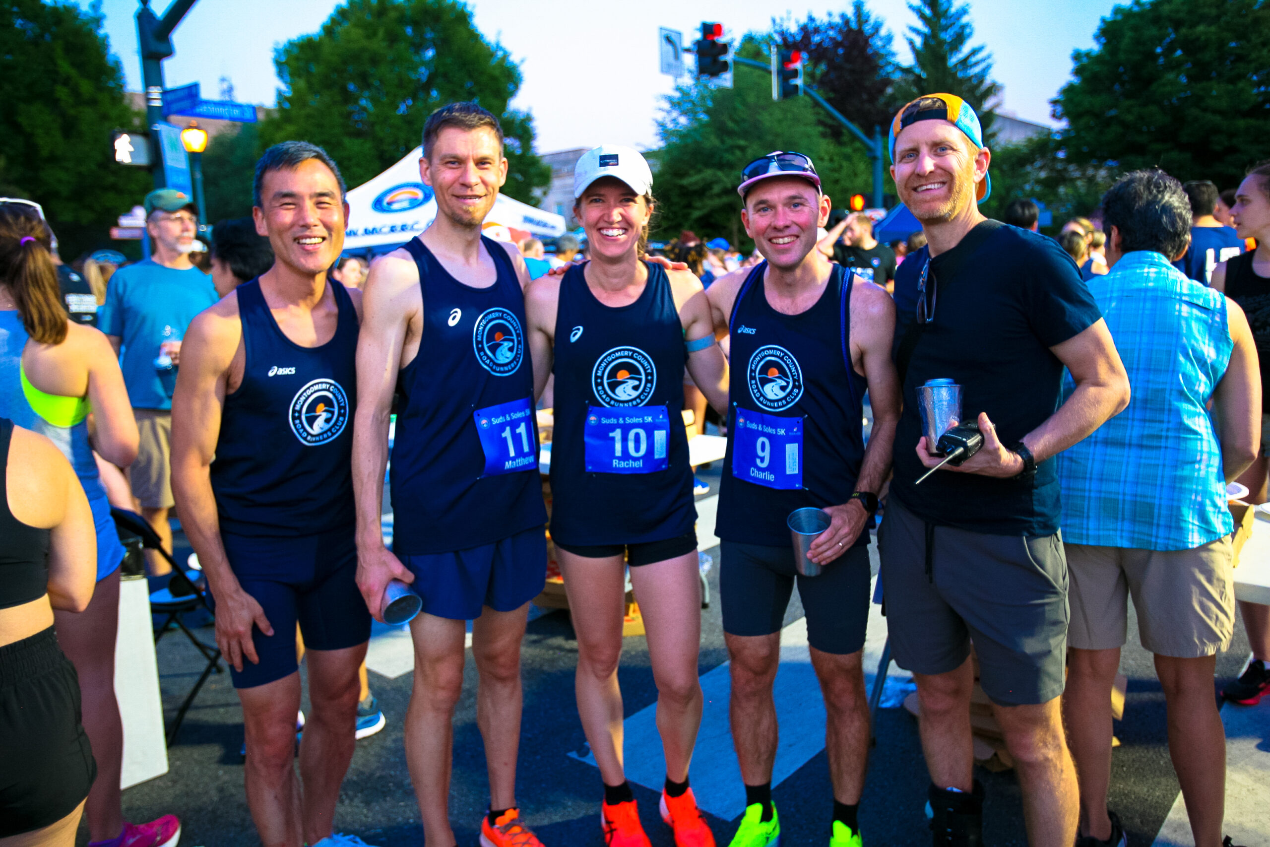 Five runners in matching team shirts and bibs pose together and smile for a photo at an outdoor race event, with other participants and trees in the background.
