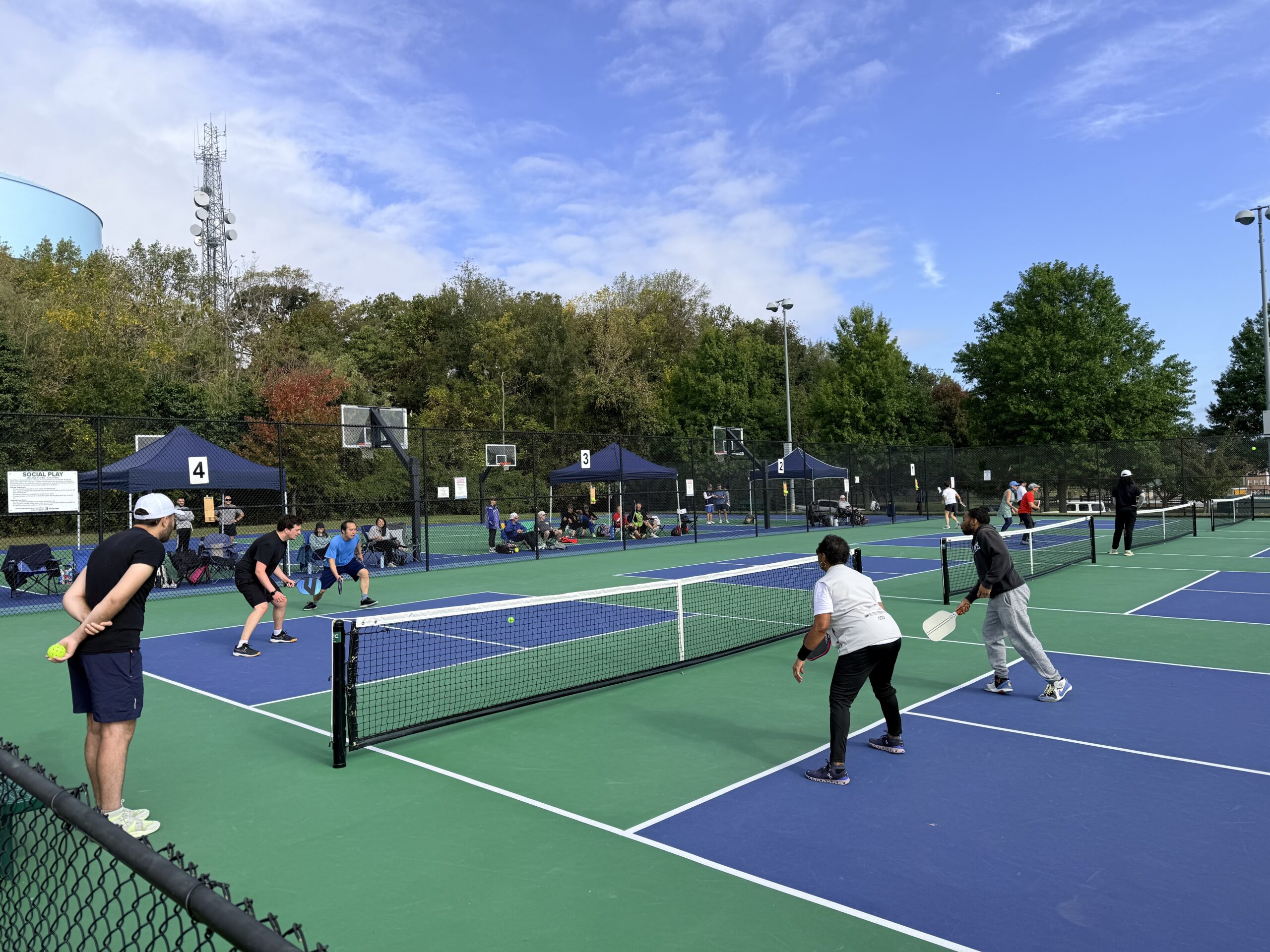 Two pairs of people play a match of pickleball on an outdoor court, with others watching and several tents in the background under a partly cloudy sky.