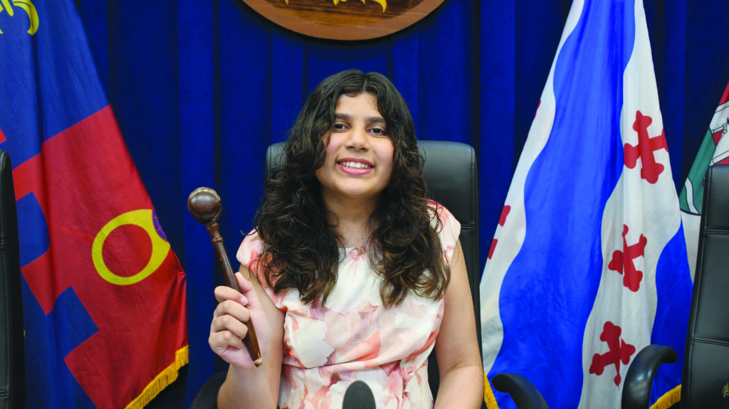 A person with long dark hair, wearing a light floral dress, sits in a large chair holding a gavel, with colorful flags in the background.