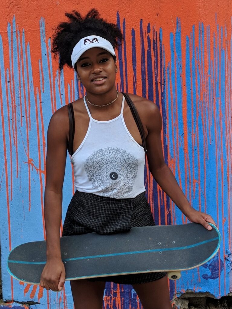 A person in a white tank top and visor holds a skateboard in front of a colorful, paint-splattered wall.