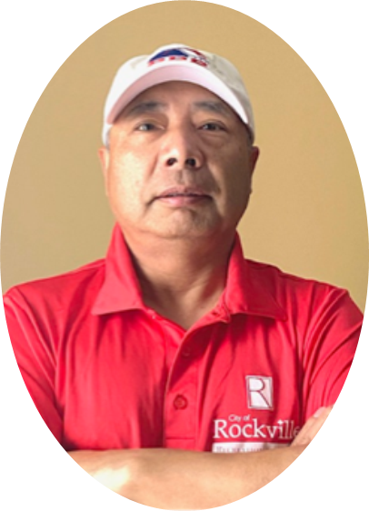 A man wearing a white cap and a red "City of Rockville" polo shirt stands with his arms crossed against a plain background.