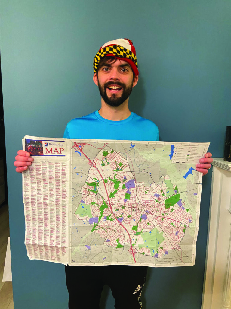 A man in a blue shirt and multicolored hat holds an unfolded map of Rockville, standing indoors against a blue wall.