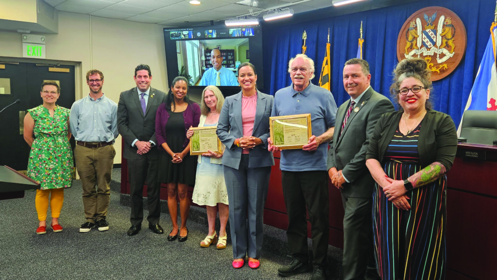A group of people, some holding framed certificates, pose for a photo in a conference room with flags and a crest in the background. A screen displays additional people joining virtually.