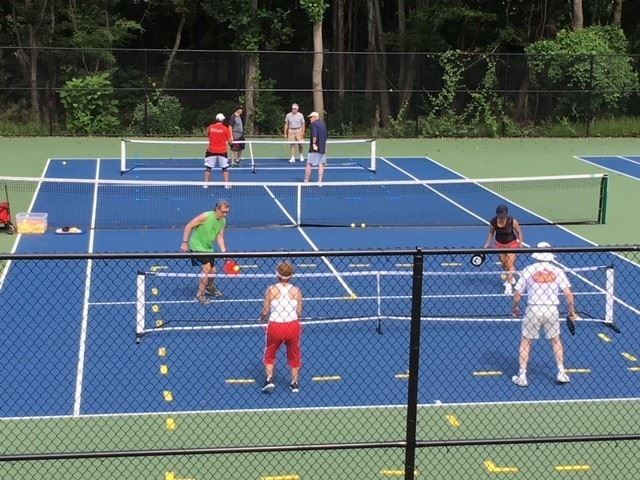 People are playing pickleball on blue outdoor courts, with two games occurring simultaneously and trees in the background.