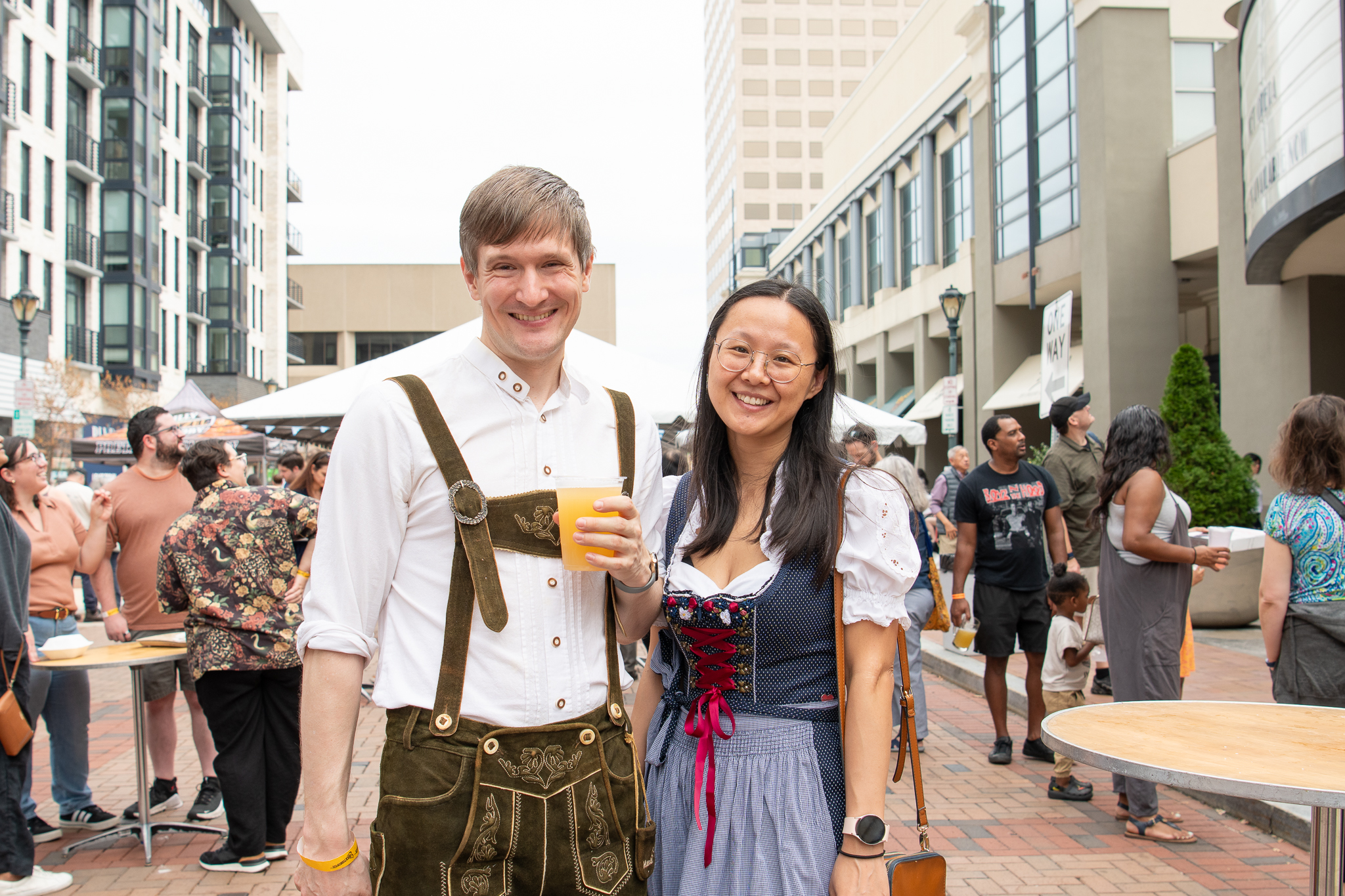 Two people in traditional Bavarian clothing stand and smile at an outdoor event with a crowd and buildings in the background. One holds a drink.