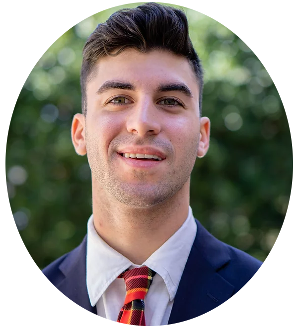 A young man in a suit and striped tie is smiling outdoors, with greenery blurred in the background.