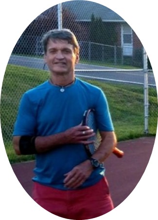Man standing on an outdoor tennis court holding a tennis racket, wearing a blue shirt, red shorts, and a black elbow support.