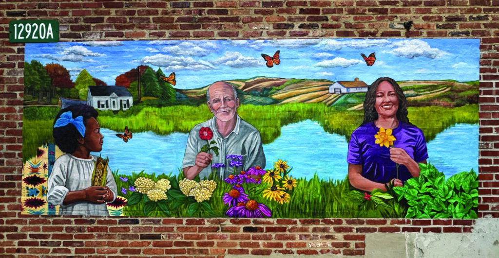 A mural on a brick wall depicts a young girl, an older man, and a woman holding flowers, with a pond, fields, houses, and butterflies in the background.