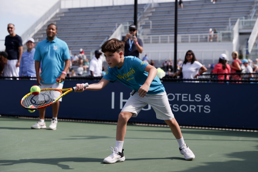 A boy in sportswear reaches to hit a tennis ball with his racket on an outdoor court, as people watch from the background.