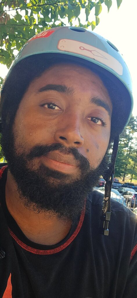 A man with a beard wearing a light blue helmet looks at the camera outdoors, with trees and parked cars in the background.