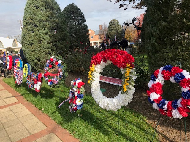 Several red, white, and blue floral wreaths are displayed on stands in a grassy area surrounded by trees during an outdoor event.
