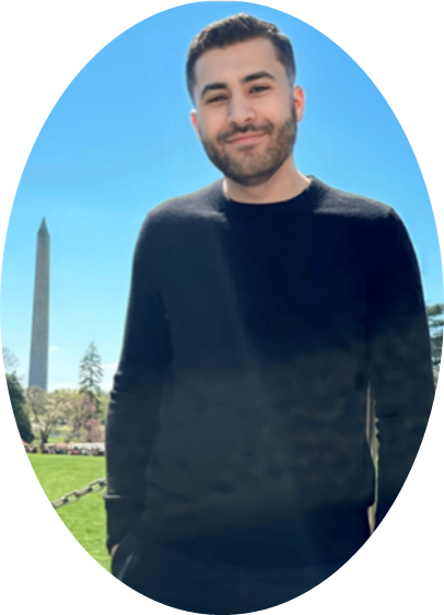 A man in a black sweater stands outdoors with the Washington Monument visible in the background on a clear, sunny day.