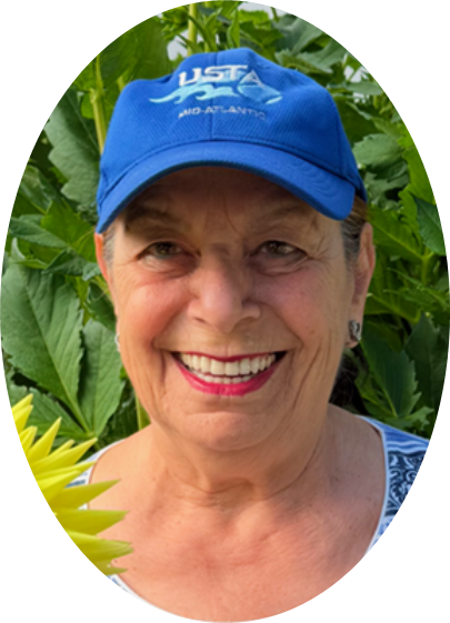 Smiling woman wearing a blue baseball cap and patterned top, standing outdoors in front of green leafy plants.
