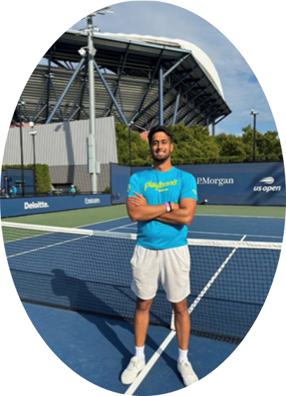 A man in a blue t-shirt and white shorts stands with arms crossed on a tennis court, with a stadium structure visible in the background.
