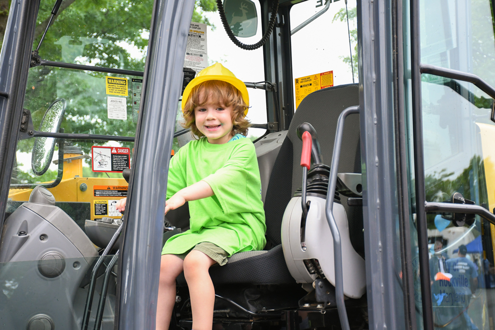 A young child wearing a yellow hard hat and green shirt sits in the driver’s seat of a construction vehicle, smiling at the camera.
