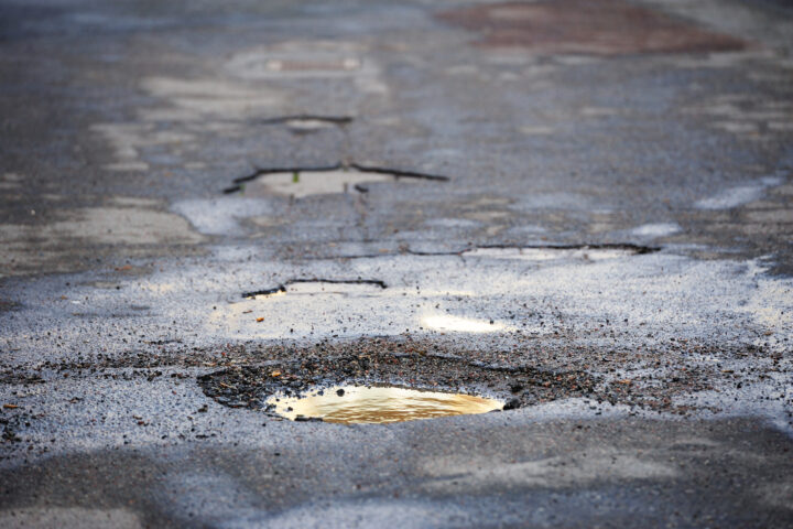 A wet asphalt road surface with several visible potholes filled with rainwater.