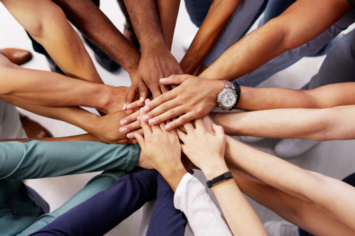 A group of people standing in a circle place their hands together in the center, showing teamwork and unity.