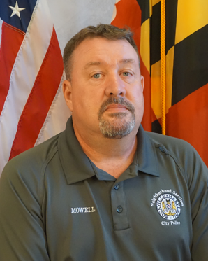 Man with short brown hair and goatee wearing a gray "City Police" polo shirt, sitting in front of U.S. and Maryland state flags.