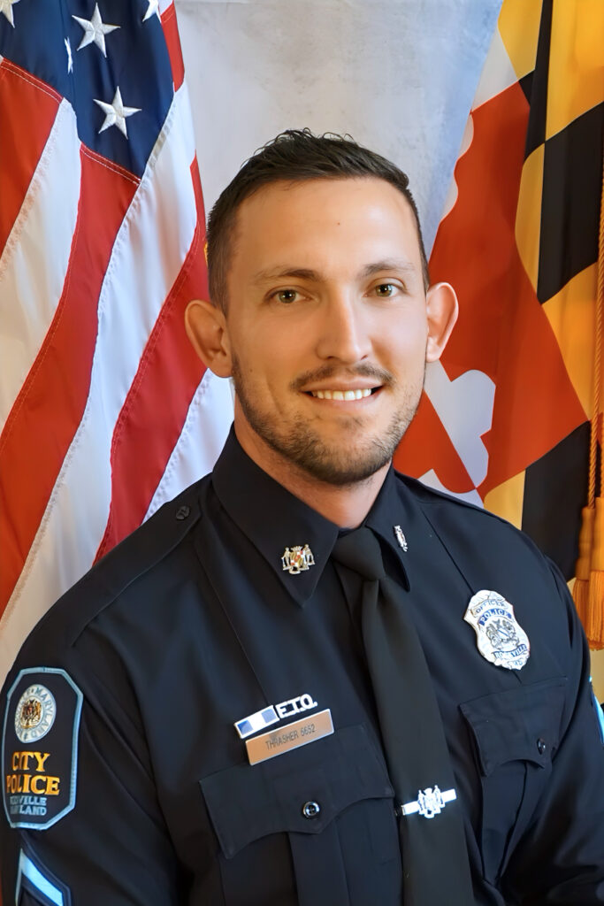 A male police officer in uniform poses for an official portrait in front of the U.S. and Maryland state flags.