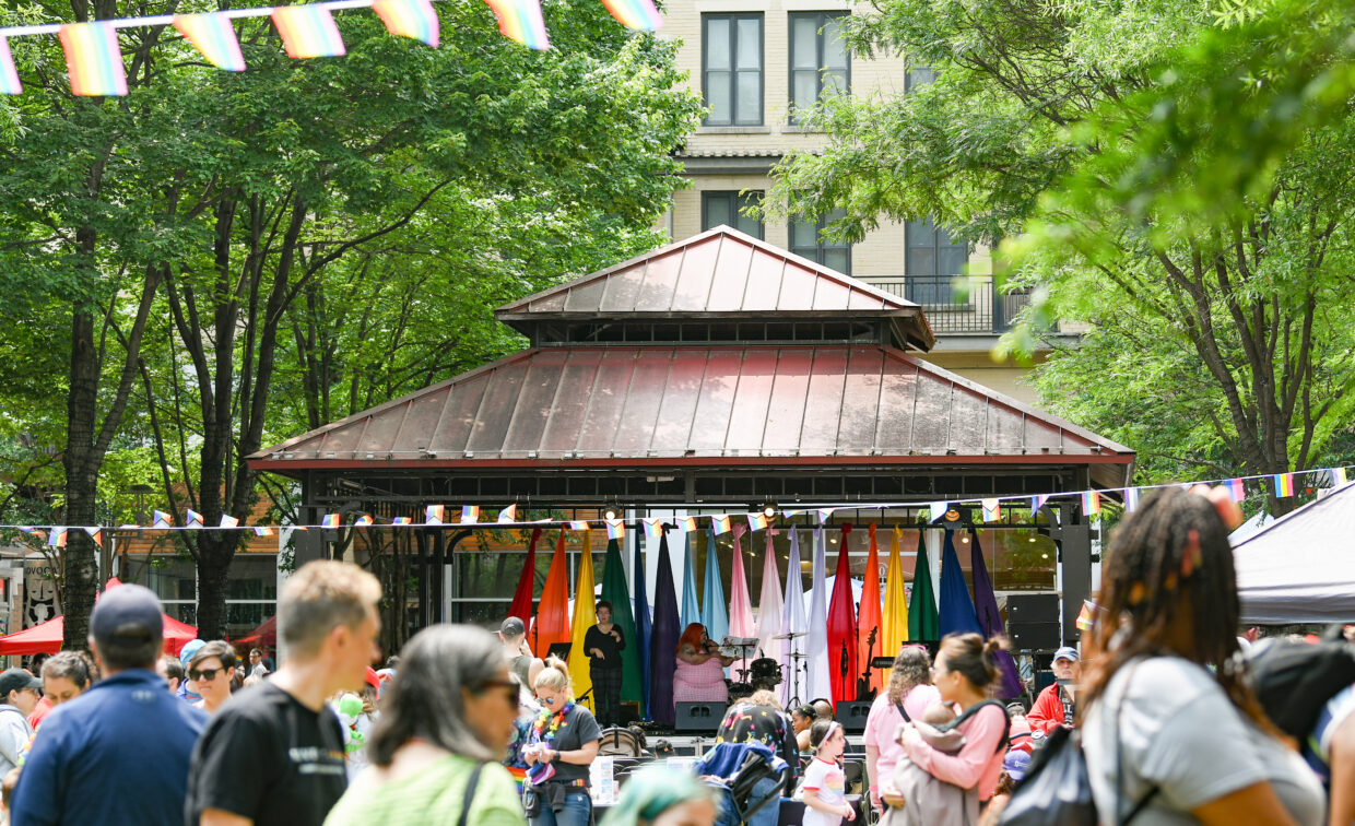 Crowd gathered in front of a gazebo decorated with international flags and rainbow banners at an outdoor event surrounded by trees.