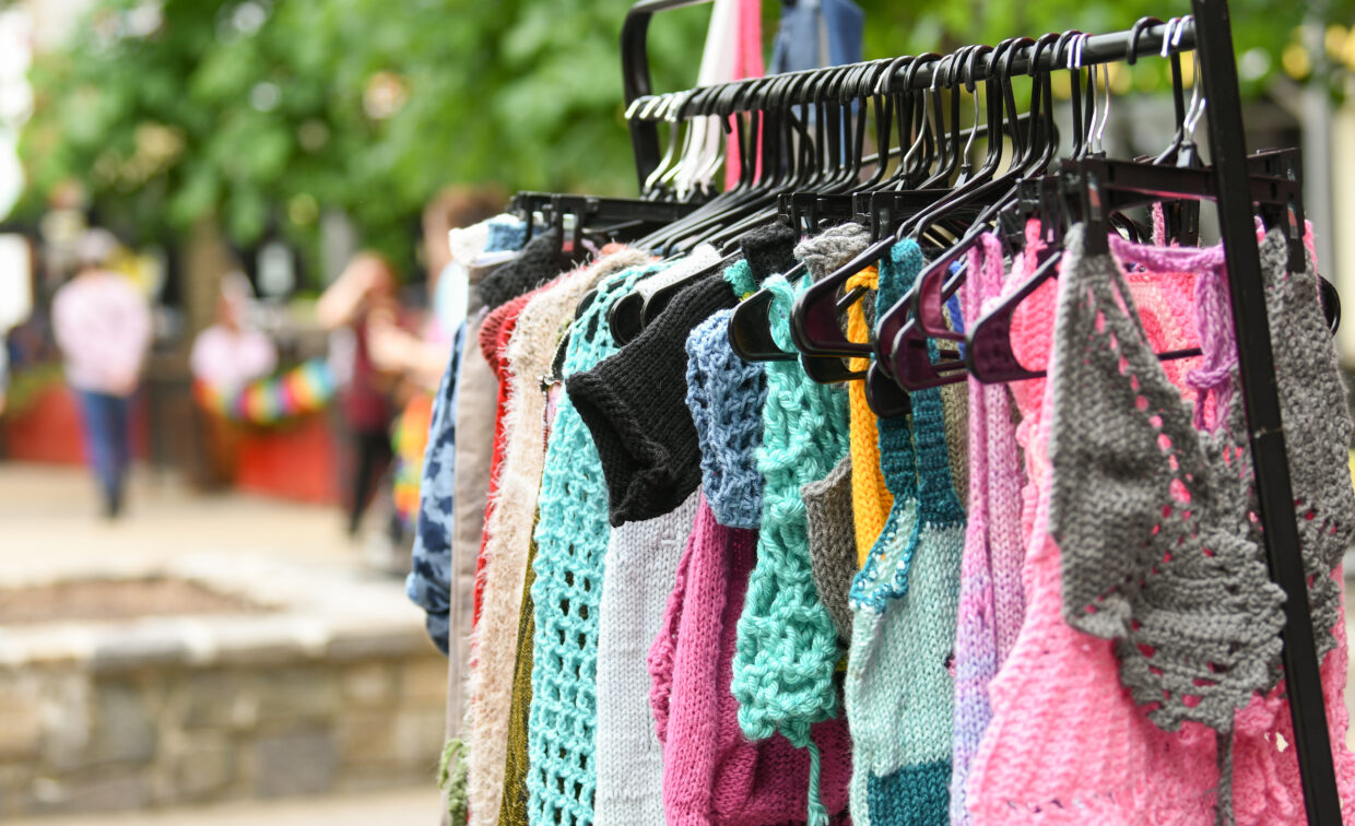 A rack displays colorful knitted and crocheted garments on hangers at an outdoor market, with trees and blurred people in the background.