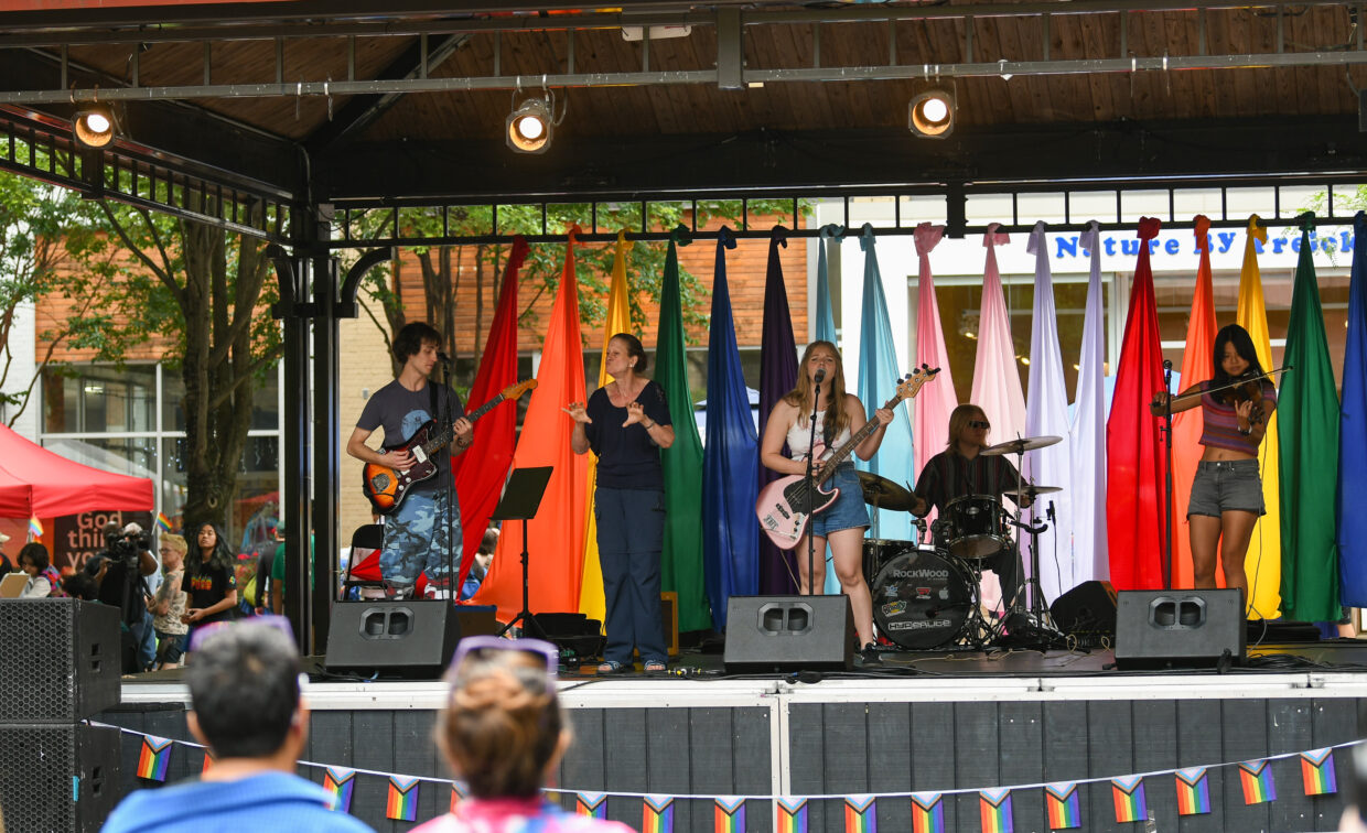 A band performs on an outdoor stage decorated with rainbow flags, while an audience watches; trees and a building are visible in the background.