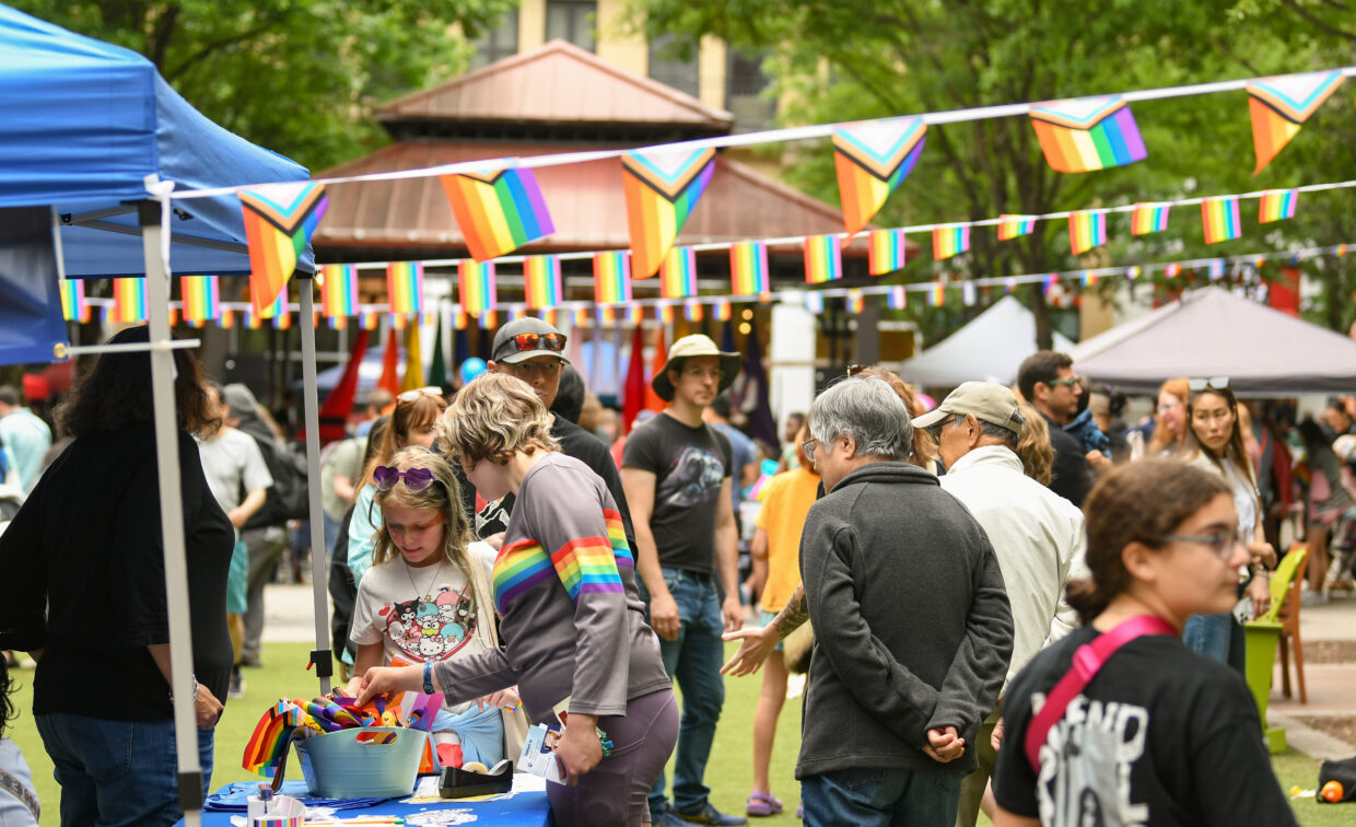 People gather at an outdoor event decorated with rainbow flags, with booths and tables set up under tents. Some attendees are interacting at a table with Pride-themed items.
