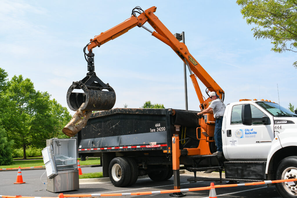 A worker operates a crane mounted on a City of Rockville truck, lifting a large metal object into a dumpster, with an old refrigerator nearby.