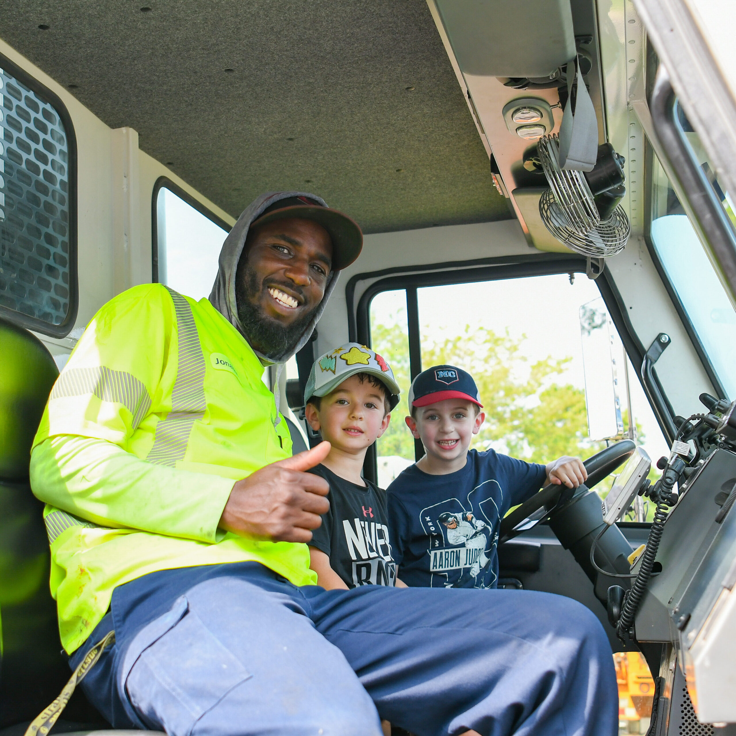 A man in a safety shirt sits in a truck cab with two young boys; one boy is in the driver’s seat holding the wheel, and the man gives a thumbs up.