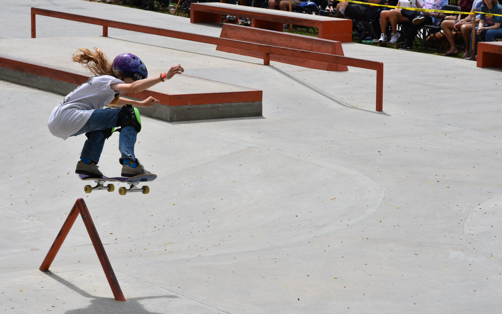 A skateboarder in mid-air performs a trick over a triangular rail at a skate park, with spectators seated in the background.