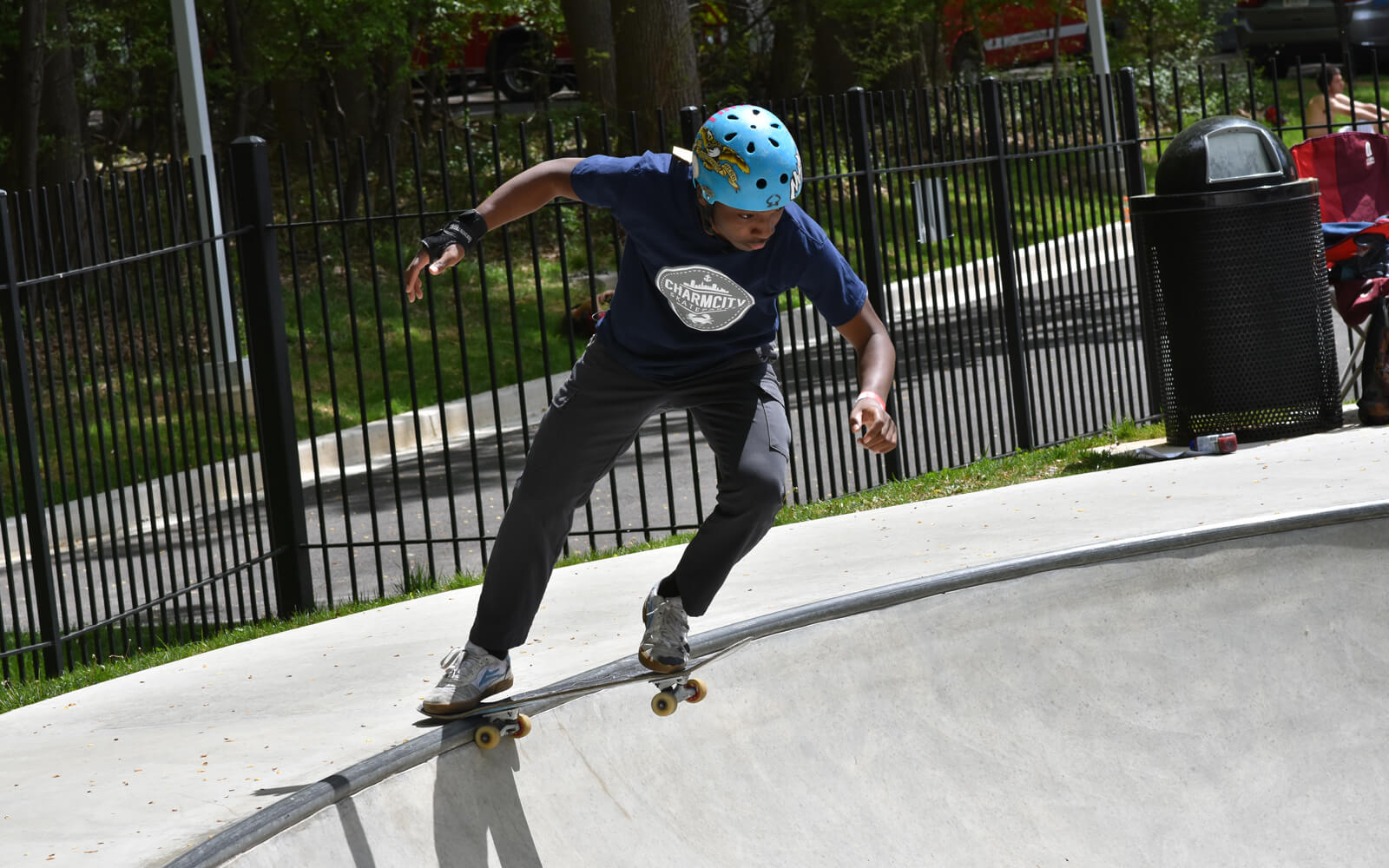 A person wearing a blue helmet and navy shirt skates on the edge of a concrete skatepark bowl, preparing to drop in.