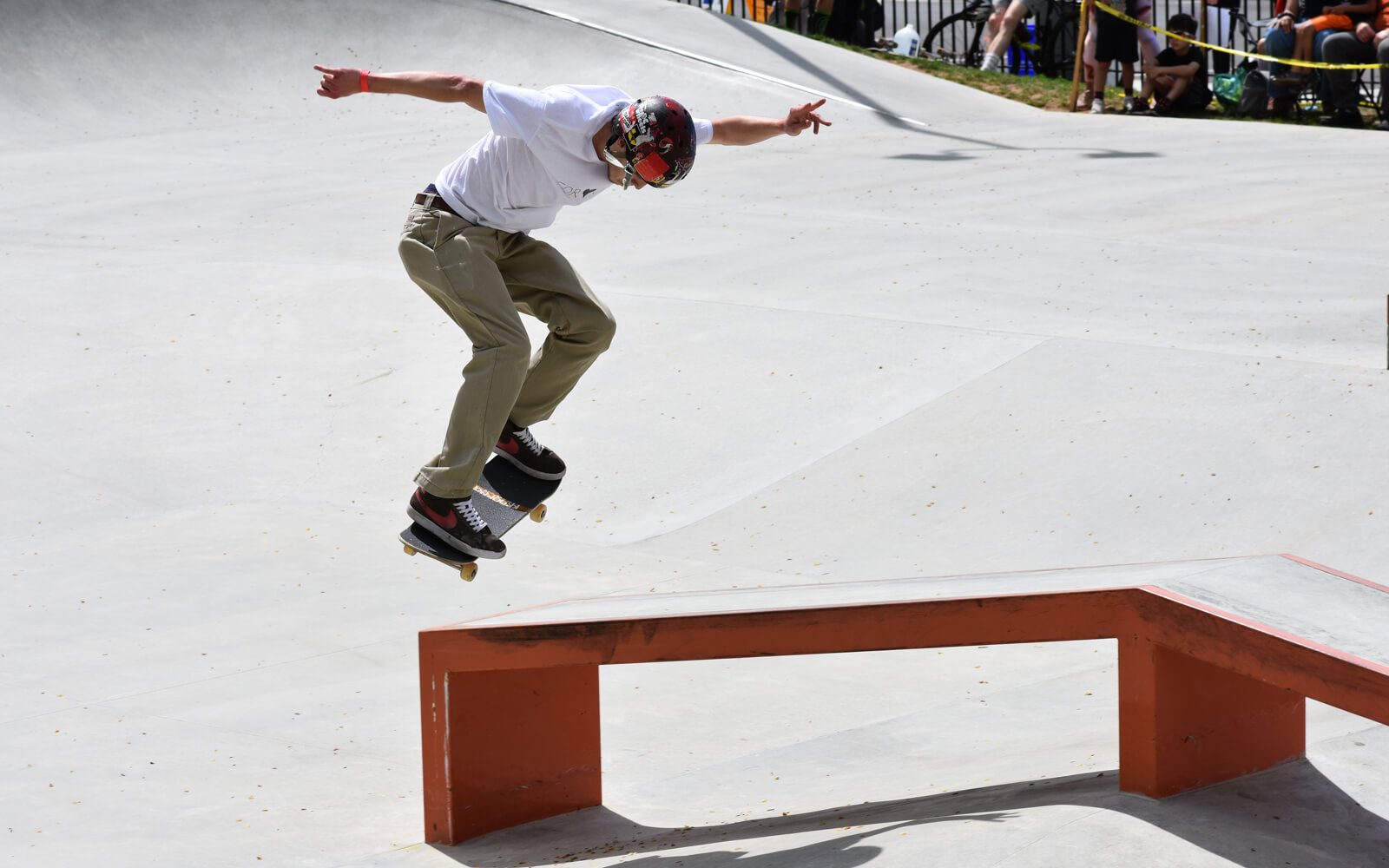 A skateboarder wearing a helmet and white shirt performs a trick over a red metal ledge at a skatepark.
