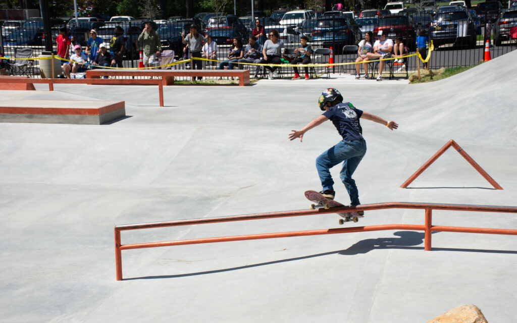 A skateboarder grinds on a rail at an outdoor skatepark while people watch from the sidelines and parked cars are visible in the background.