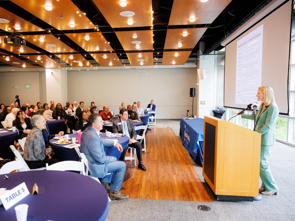 Maryland Comptroller Brooke Lierman speaks at a podium to an audience seated at round tables in a modern conference room with wood-accented ceiling and bright lighting.