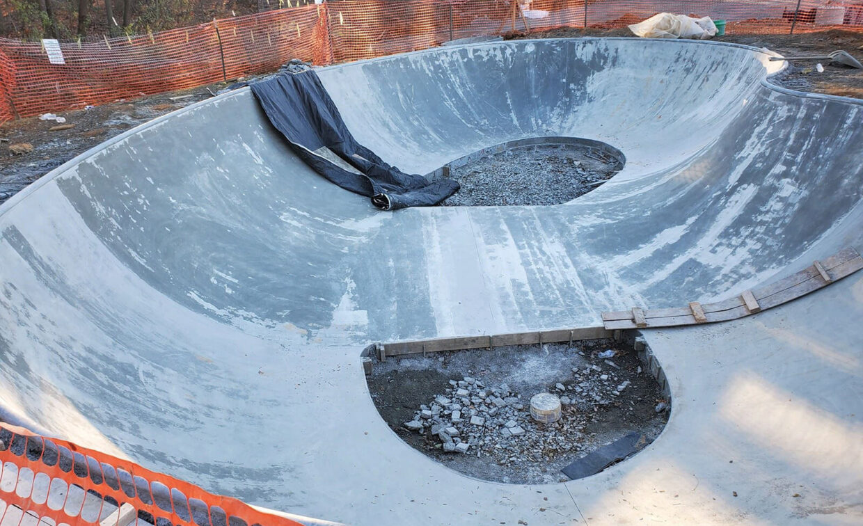 Large concrete skate bowl under construction with unfinished sections, surrounded by orange safety fencing and construction materials.