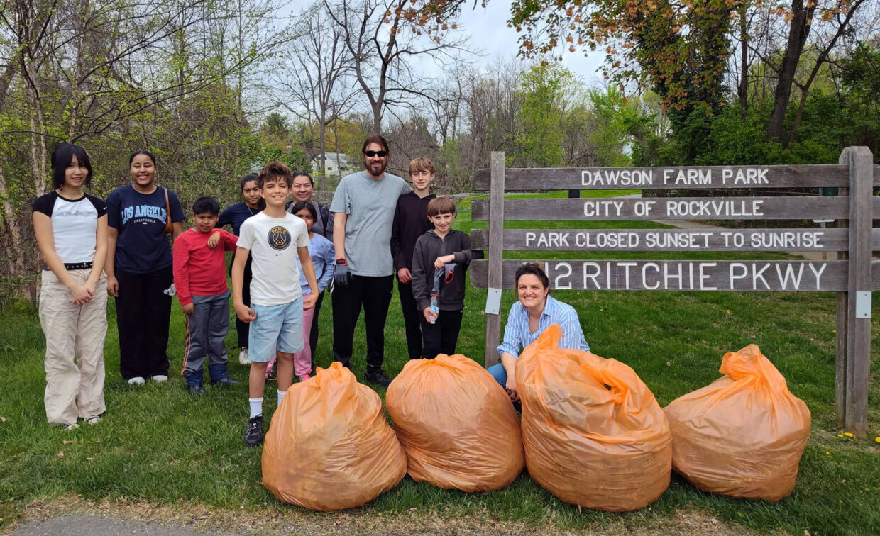 A group of people pose by large orange trash bags in front of a Dawson Farm Park sign, after collecting litter. Trees and grass are visible in the background.