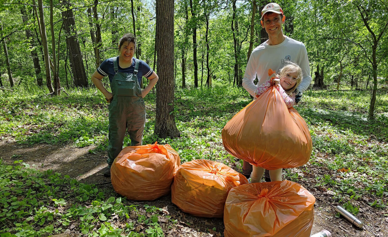 Two adults and a child stand in a wooded area with four large orange trash bags filled with collected litter.