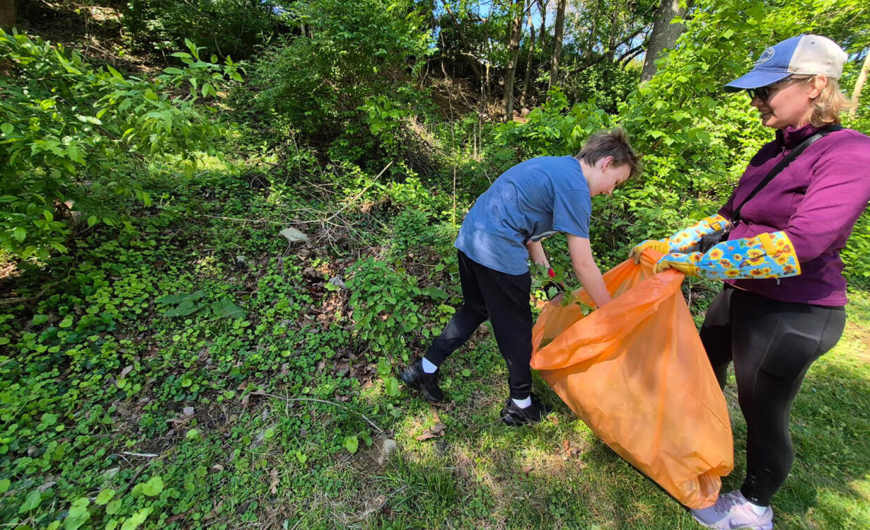 A woman and a child pick up litter and place it in an orange bag while standing on grass near dense green foliage.