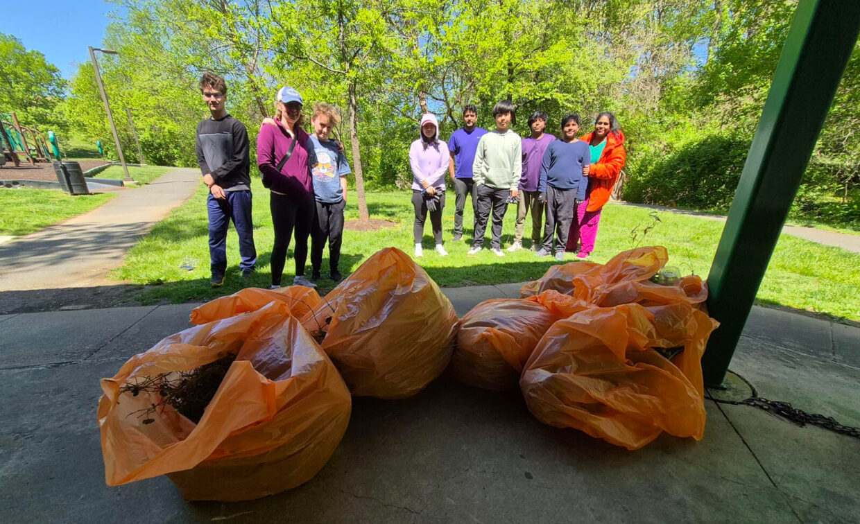 A group of people stands outdoors on a sunny day behind large orange bags filled with collected yard waste or debris.