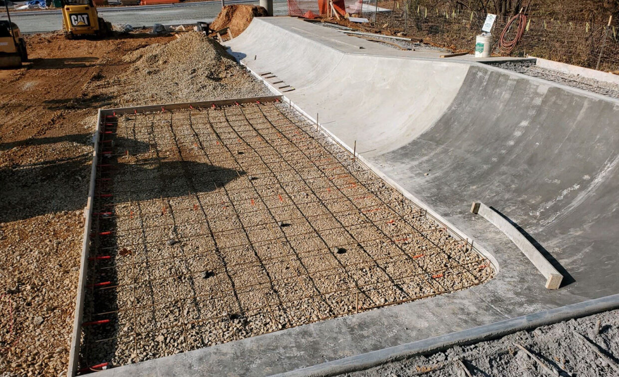 Concrete skatepark under construction showing a partially completed ramp and a section with exposed rebar grid over gravel, ready for pouring concrete. Construction equipment visible in background.