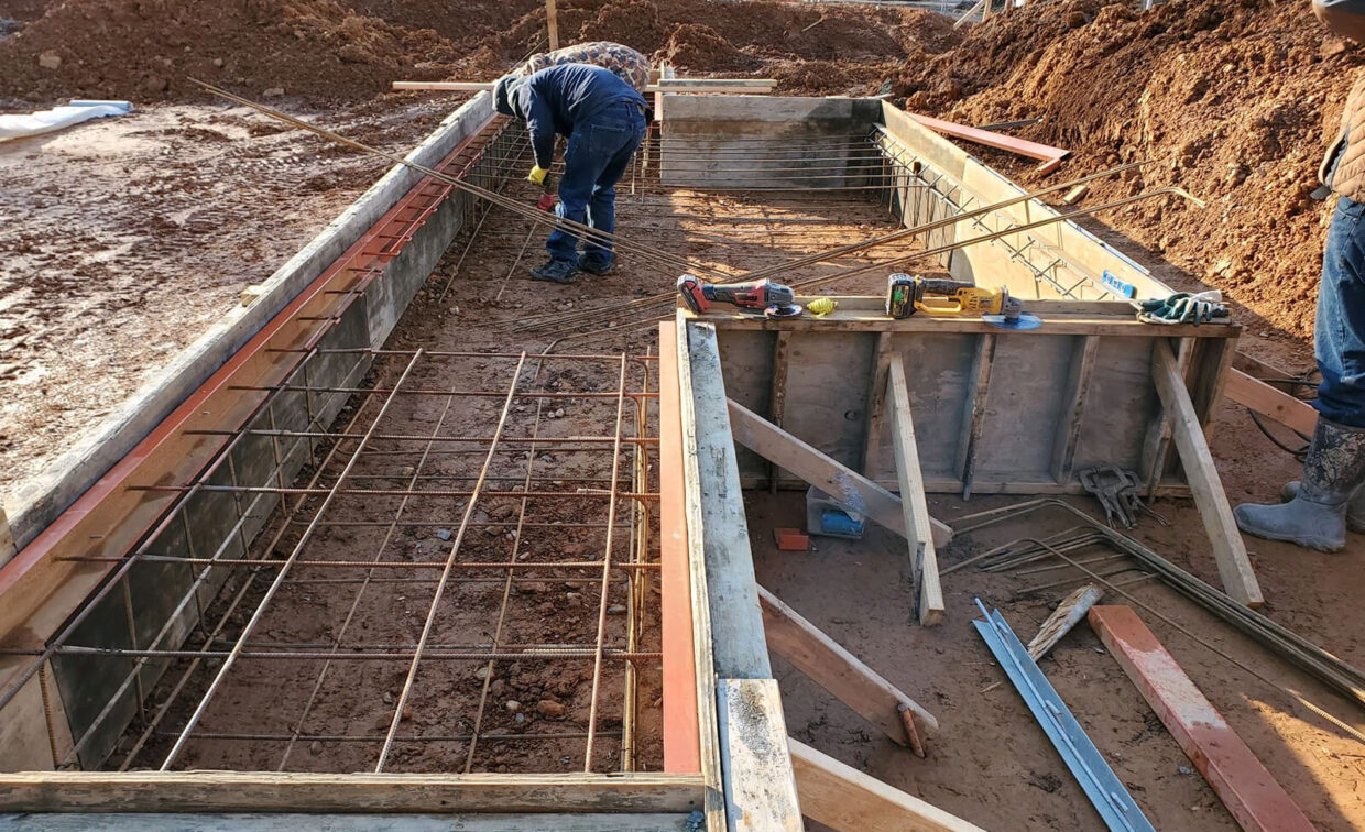 Construction workers preparing rebar and wooden forms for a concrete foundation at a building site with tools and materials scattered nearby.