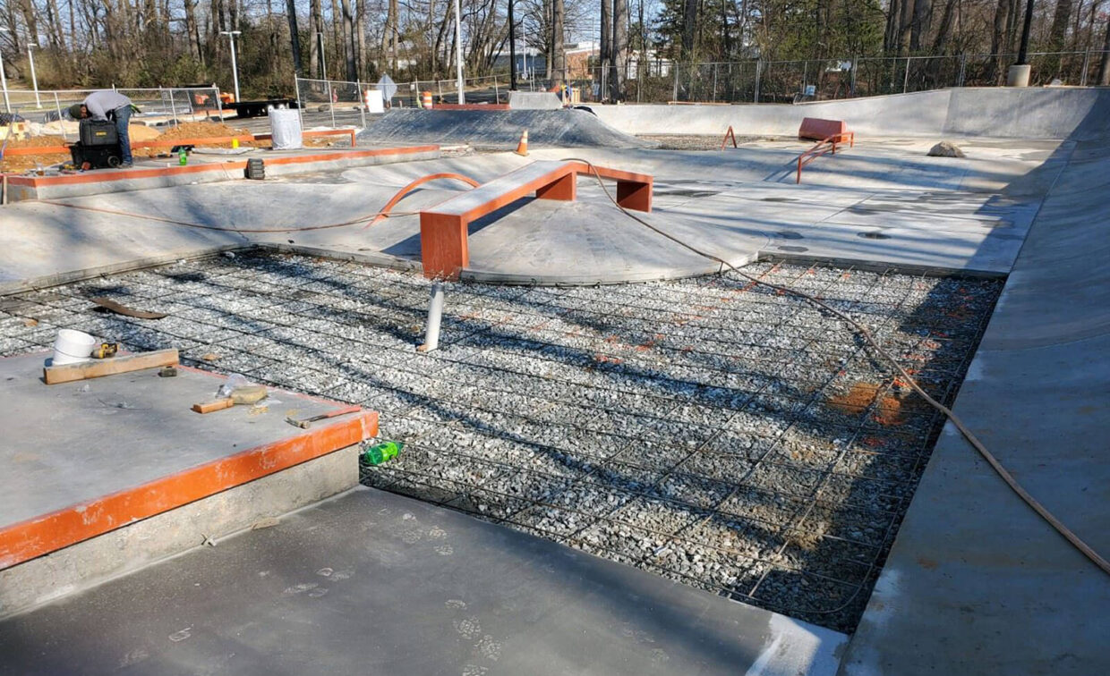 A concrete skatepark under construction with exposed rebar, gravel, and partially built ramps and features.
