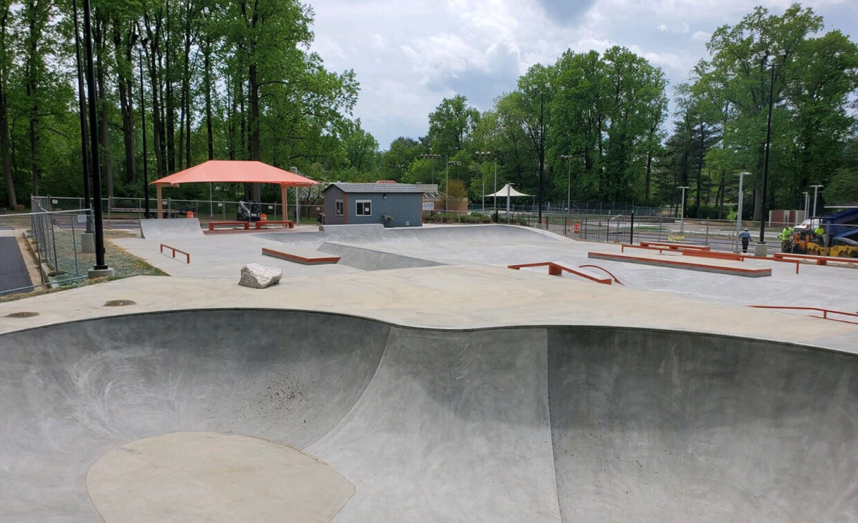 A concrete skatepark with ramps, rails, and bowls, surrounded by trees and a fence, with a small building and shade structures in the background.