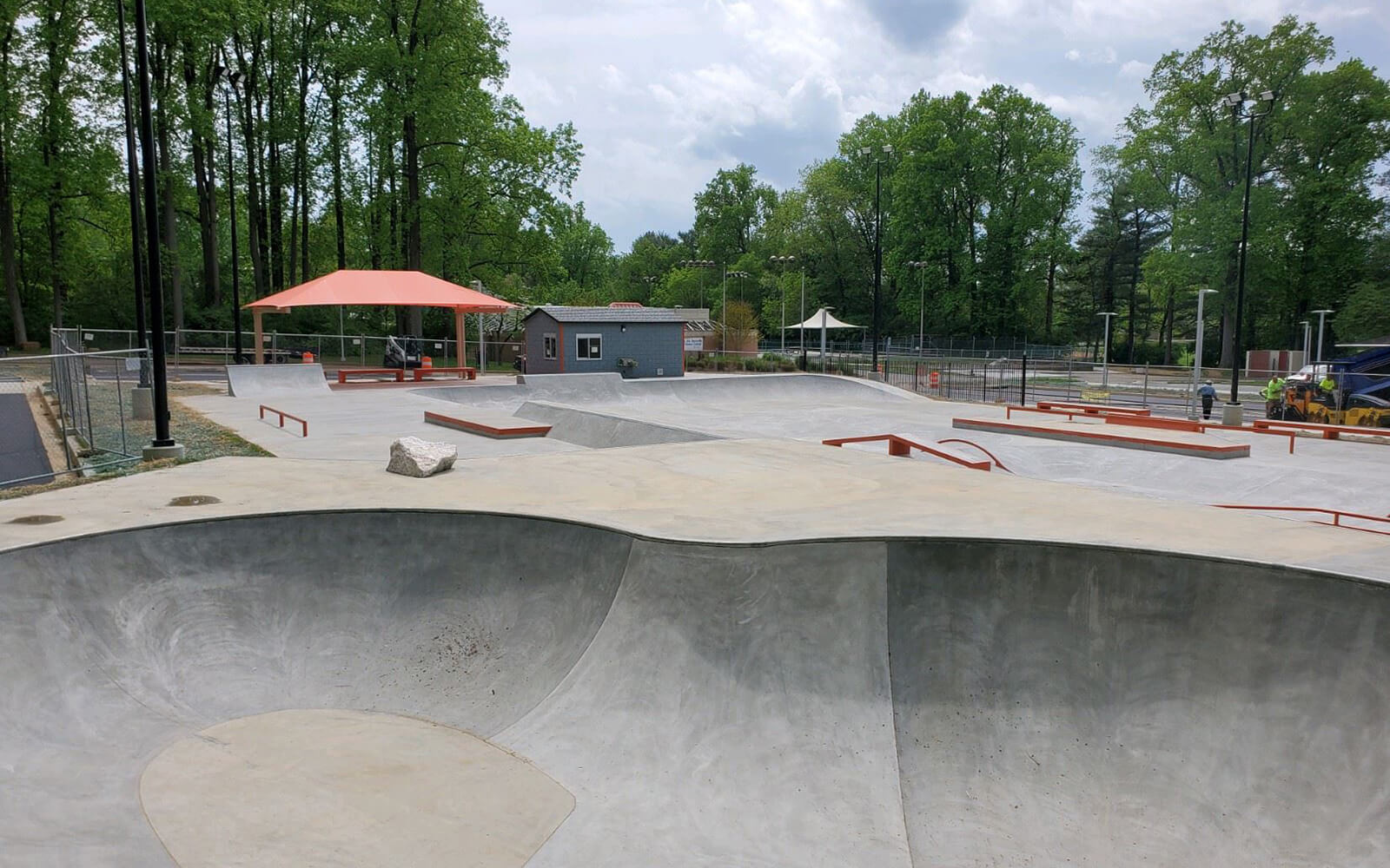 A concrete skatepark with ramps, rails, and bowls, surrounded by trees and a fence, with a small building and shade structures in the background.
